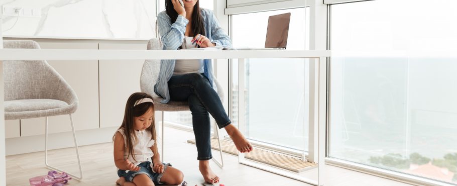 Young asian woman talking on a mobile phone and working on laptop while her little daughter playing on a floor at home